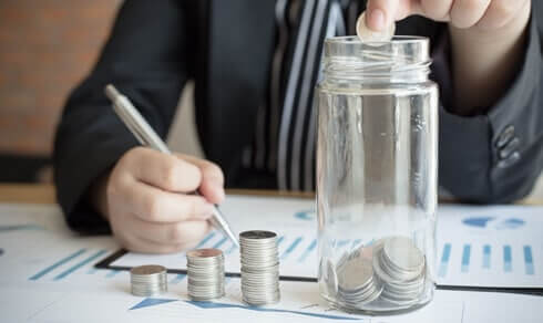 A person putting a coin in a jar and writing with other hand - Greenberg Financial Group